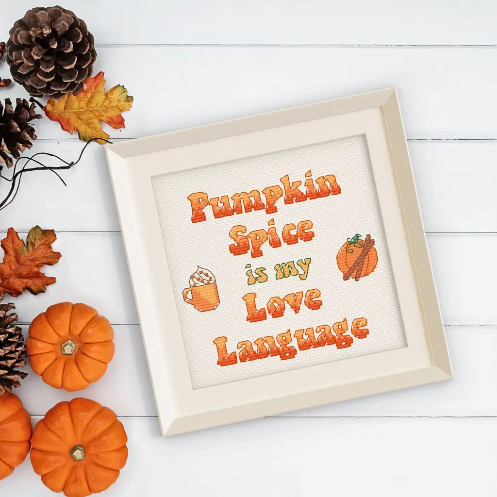 Finished Pumpkin Spice Lover cross stitch by Room for Calm in a cream colored frame on a table with pumpkins, fall leaves, and pinecones