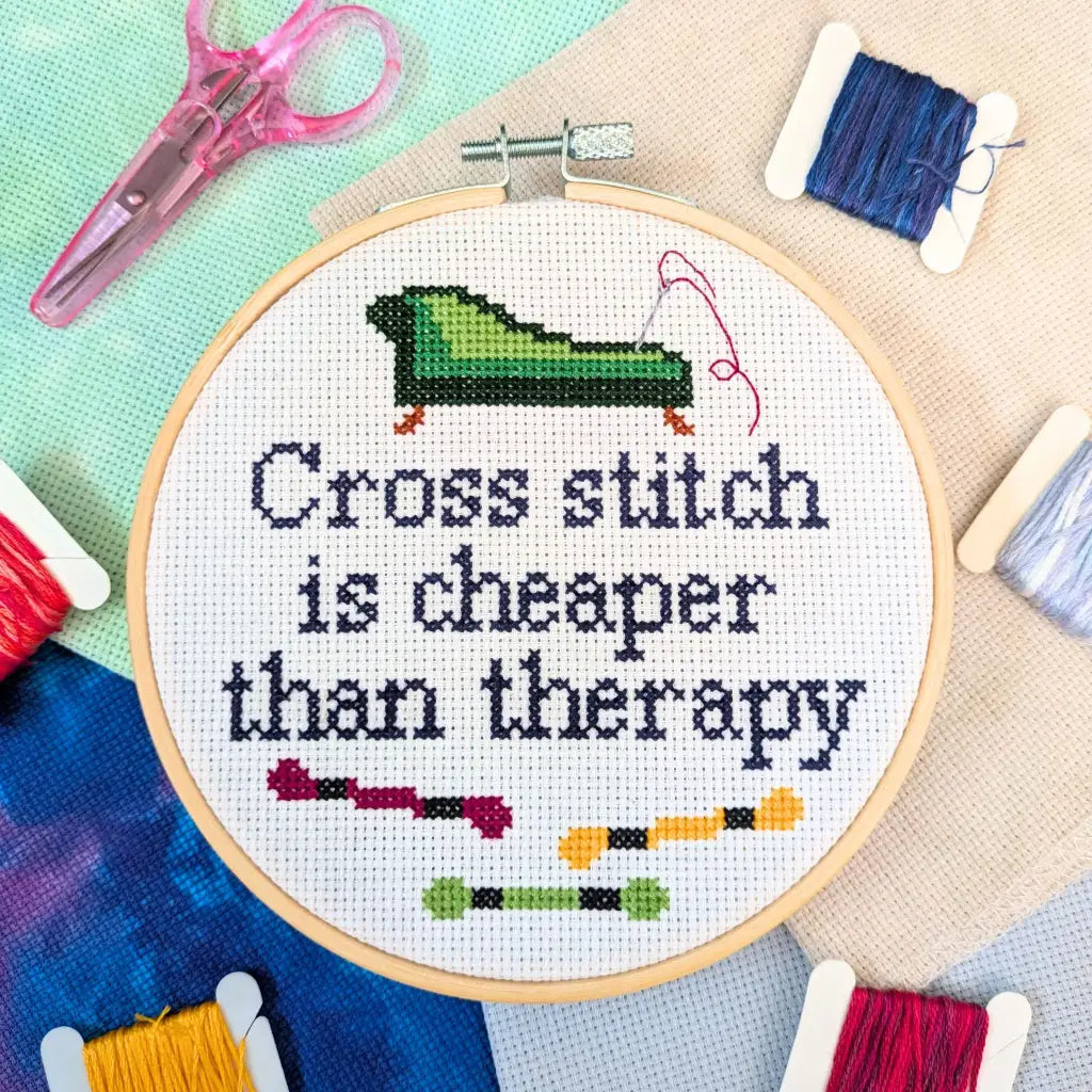 Cross stitch hoop with 'Cross stitch is cheaper than therapy' text and embroidery tools on a colorful background