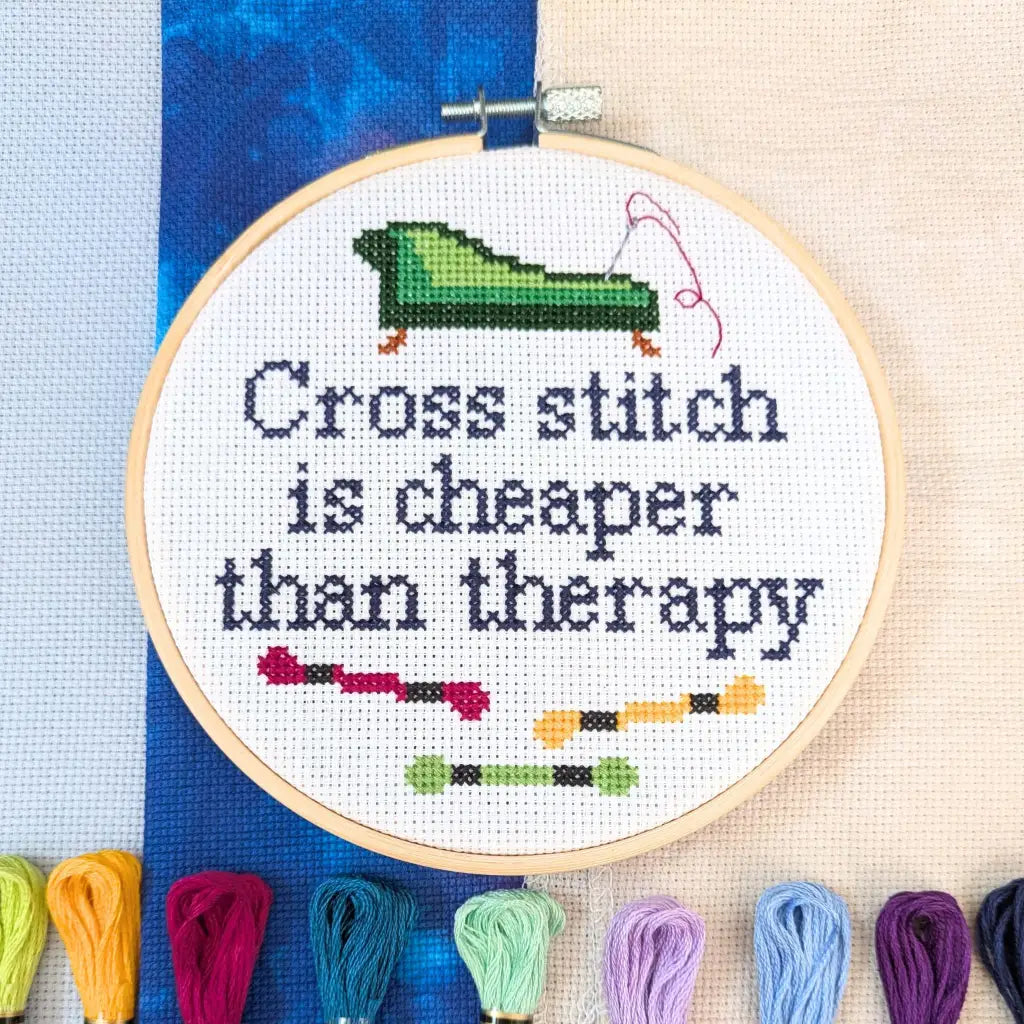 Cross stitch hoop with 'Cross stitch is cheaper than therapy' text and colorful threads on a blue fabric background.