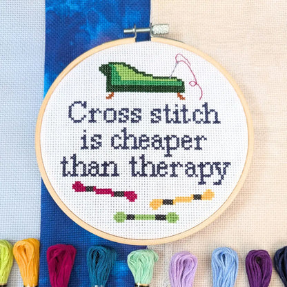 Cross stitch hoop with 'Cross stitch is cheaper than therapy' text and colorful threads on a blue fabric background.