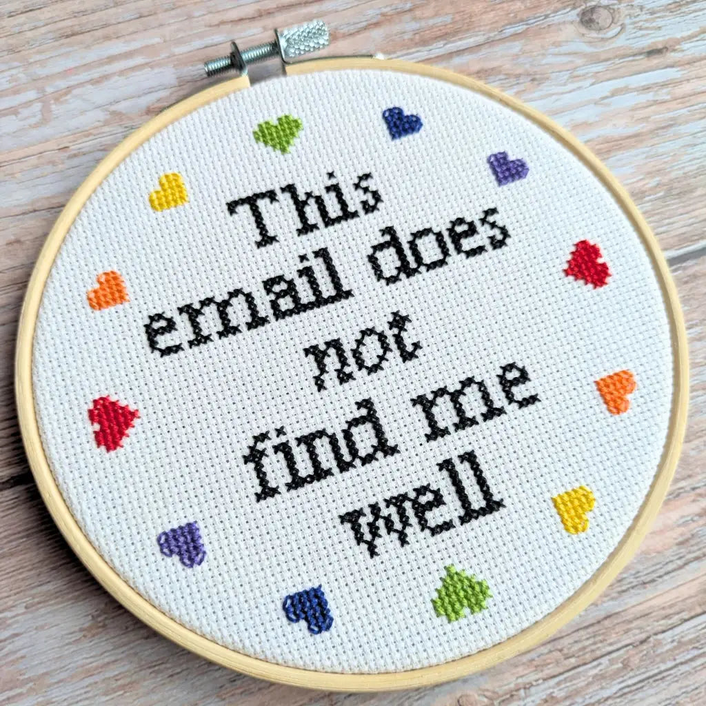 Embroidery hoop with cross-stitched text 'This email does not find me well' on a wooden surface.