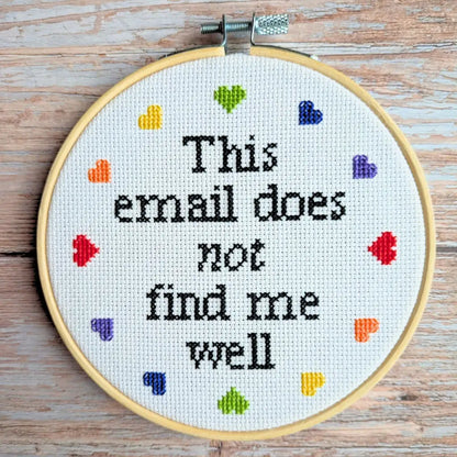 Embroidery hoop with cross-stitched text 'This email does not find me well' on a wooden surface.